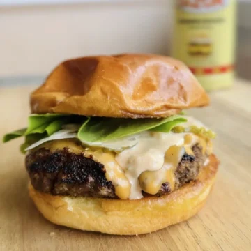 Premium beef burger patty topped with melted cheese, creamy sauce, and fresh arugula on toasted brioche bun for foodservice distribution.