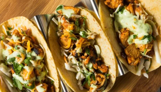 Three plated chicken tacos with crispy meat, creamy sauce, shredded cabbage, and cilantro garnish on wooden surface.
