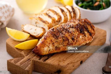 Roasted chicken breast with lemon wedges and sautéed greens on a rustic wooden board.