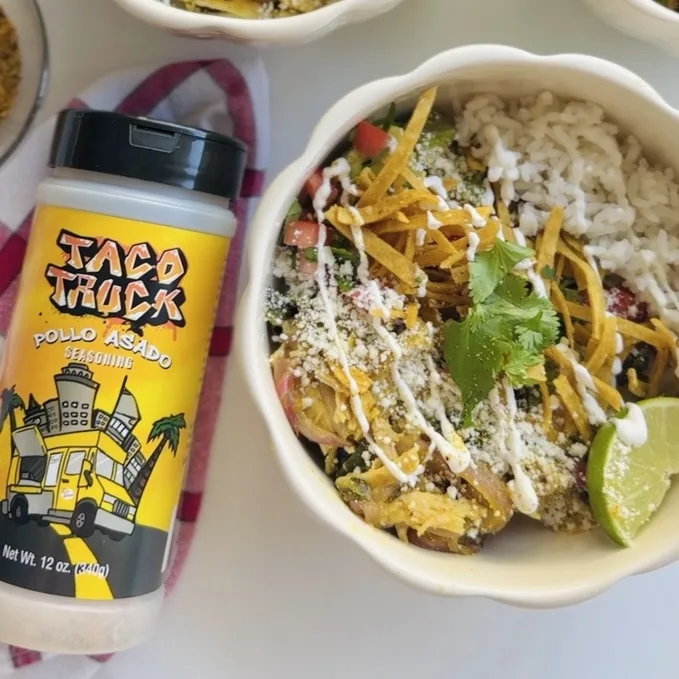 Plated pollo asado rice bowl with crispy tortilla strips, fresh cilantro, queso fresco, and lime wedge beside Taco Truck branded seasoning bottle.